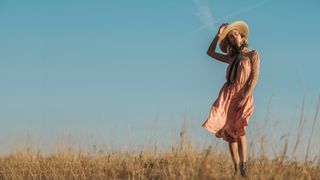 A woman standing in a field wearing a sundress and sun hat, the wind is blowing her dress gently