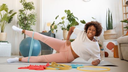 A woman exercises on a yoga mat with resistance bands. She is lying on her side, propped up on her forearm, with a band looped round her lower legs. She is raising her top leg so that it's parallel with the ground and smiling at the camera. Behind her are several leafy plants and a couch.