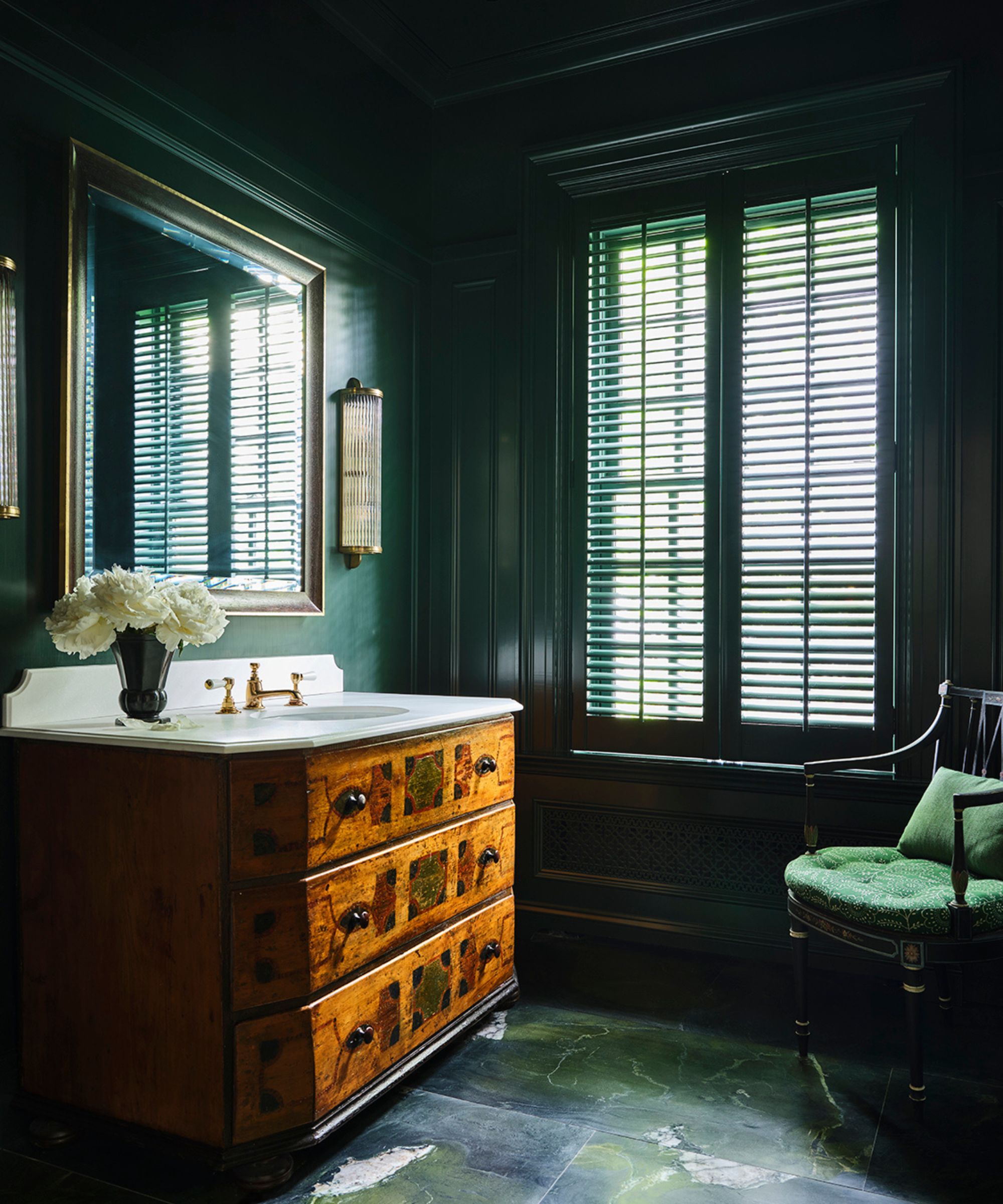 A moody, luxurious bathroom with dark emerald green walls, green stone flooring, and a window with slated blinds. A large wooden dresser with a marble countertop and sink, brass hardware, a large mirror with brass and glass sconces, and a green accent chair.
