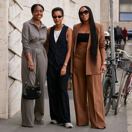 Three Black women pose together for a street style photo. One is wearing a gray set. One is wearing a black vest set. One person is wearing a brown suit. 