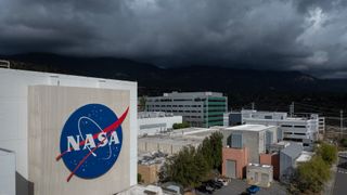 a white building with a round blue NASA logo takes up the foreground, while a storm clouds can be seen in the distance above a mountain range in the background