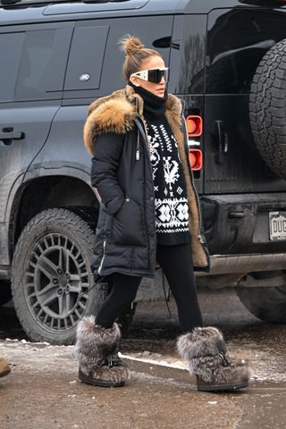 Jennifer Lopez wears large sunglasses, a black turtleneck sweater, a black fur-lined winter coat, and black leggings.