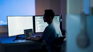 Cyber security analyst using AI security tools at desktop workstation in low-lit room.