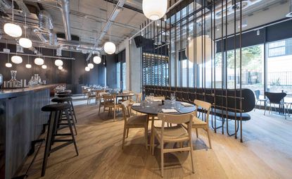 Dining space at Borealis restaurant with decorated tables, light wood chairs, wooden bar counter, dark bar chairs, round pendant lights, wooden floors and large windows.