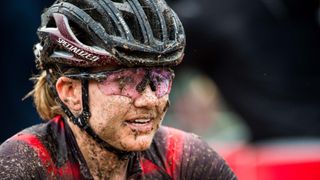 A close up shot of a woman wearing a Specialized gravel helmet and sunglasses, completely covered in mud splatters after a cyclocross race