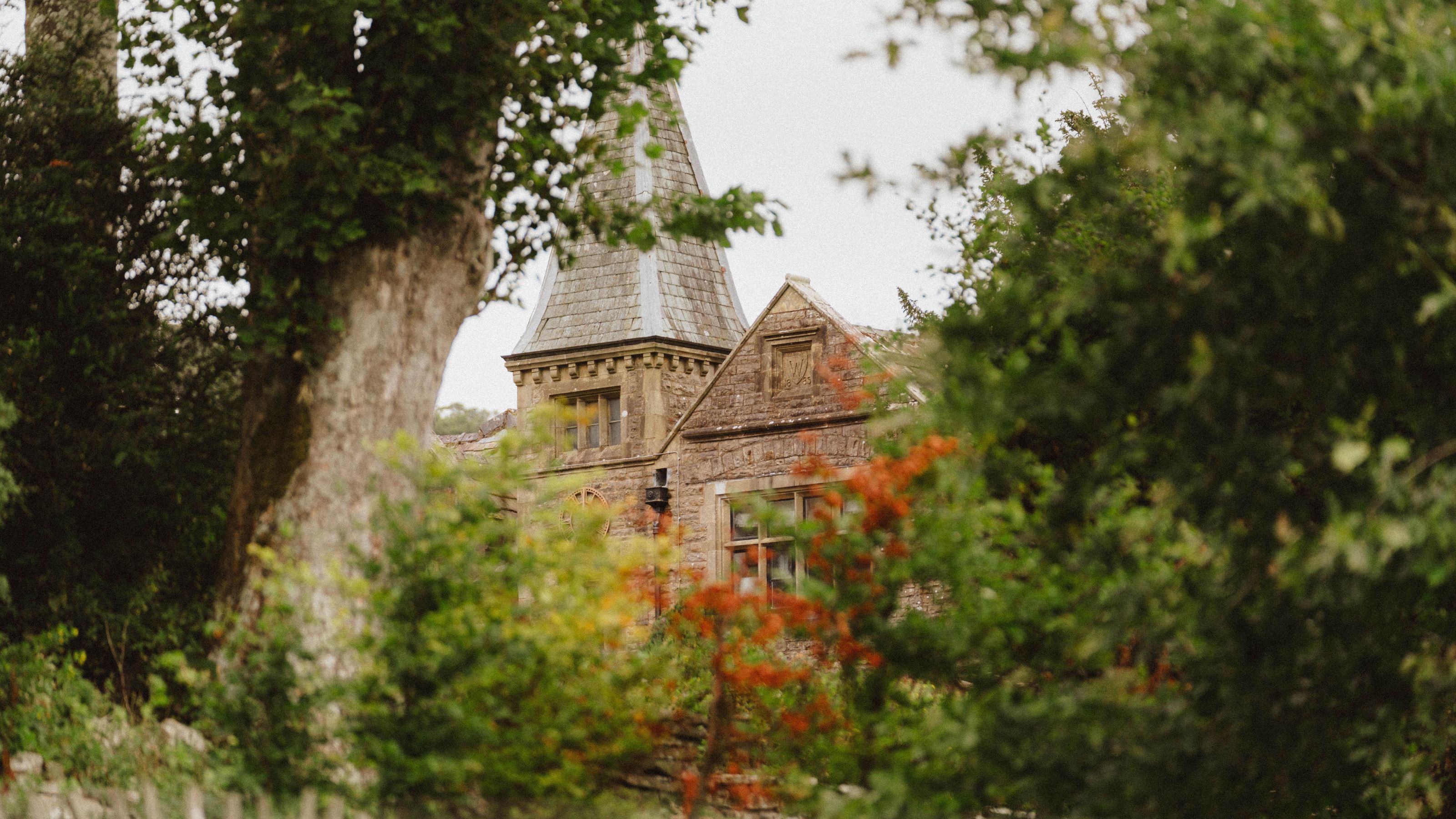 A leafy, autumnal view of a brick wall, historical country lodge, surrounded on either hand by vegetation.