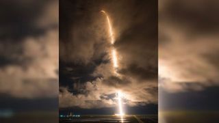 A streak of light shoots in a tall arc from the horizon, rising in the sky, through dimly lit cloud cover.