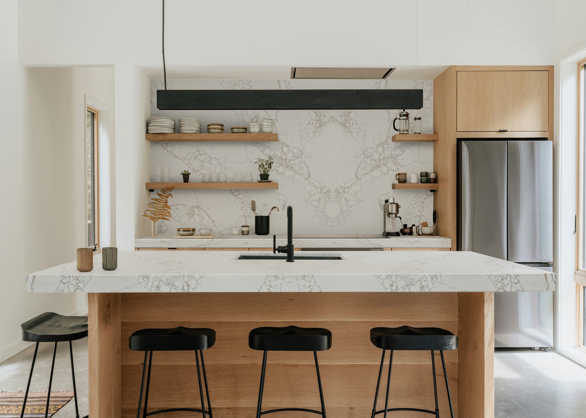 A view of a large kitchen island with black bar stools beside it and a hanging light. The countertop leads to the backsplash, all the way to the ceiling. There is also open shelving with kitchen essentials.