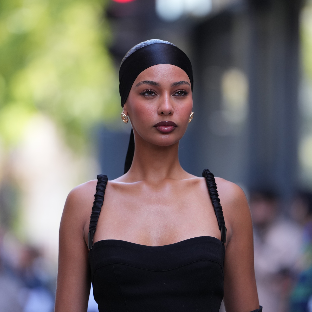 Treatment stacking Jamilla Strand wears a black satin headscarf tied at the nape with a trailing tail, gold earrings, a form-fitting black midi dress in a smooth fabric with braided spaghetti straps, a sweetheart neckline and a high thigh slit, elbow-length sheer black gloves, sheer black tights, outside Mugler, during Paris Fashion Week 