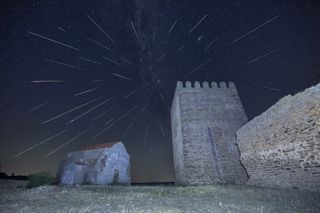 A castle-like tower and wall stand on the right, next to a small, stone building. Streaks of shooting stars stretch toward you from the night sky.