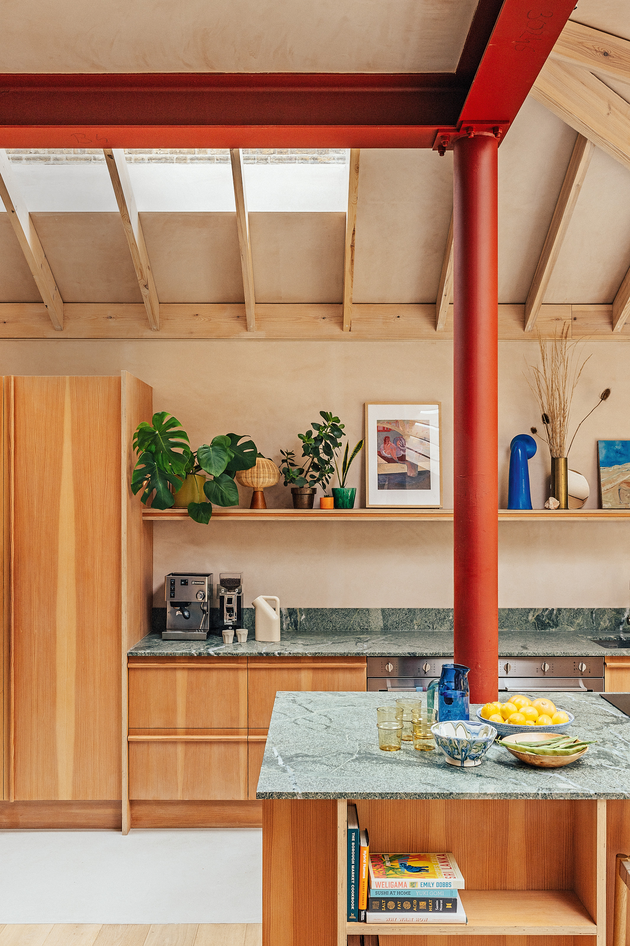 Kitchen with exposed red structural steel, wood cabinetry with green stone worktop, plaster pink walls and exposed roof rafters