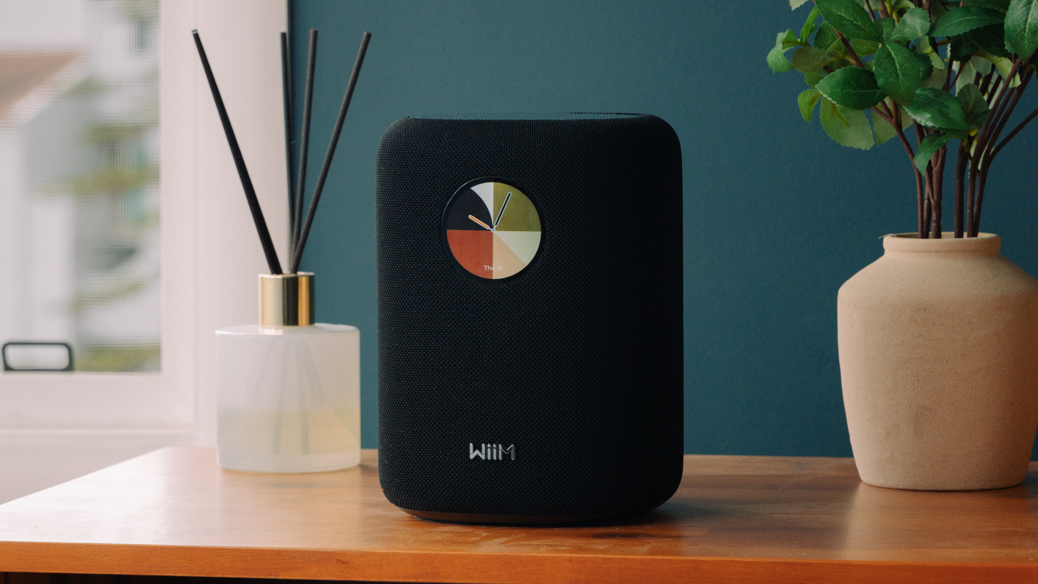 WiiM Sound speaker in black atop a wooden unit. It's flanked by a reed diffuser and a small plant
