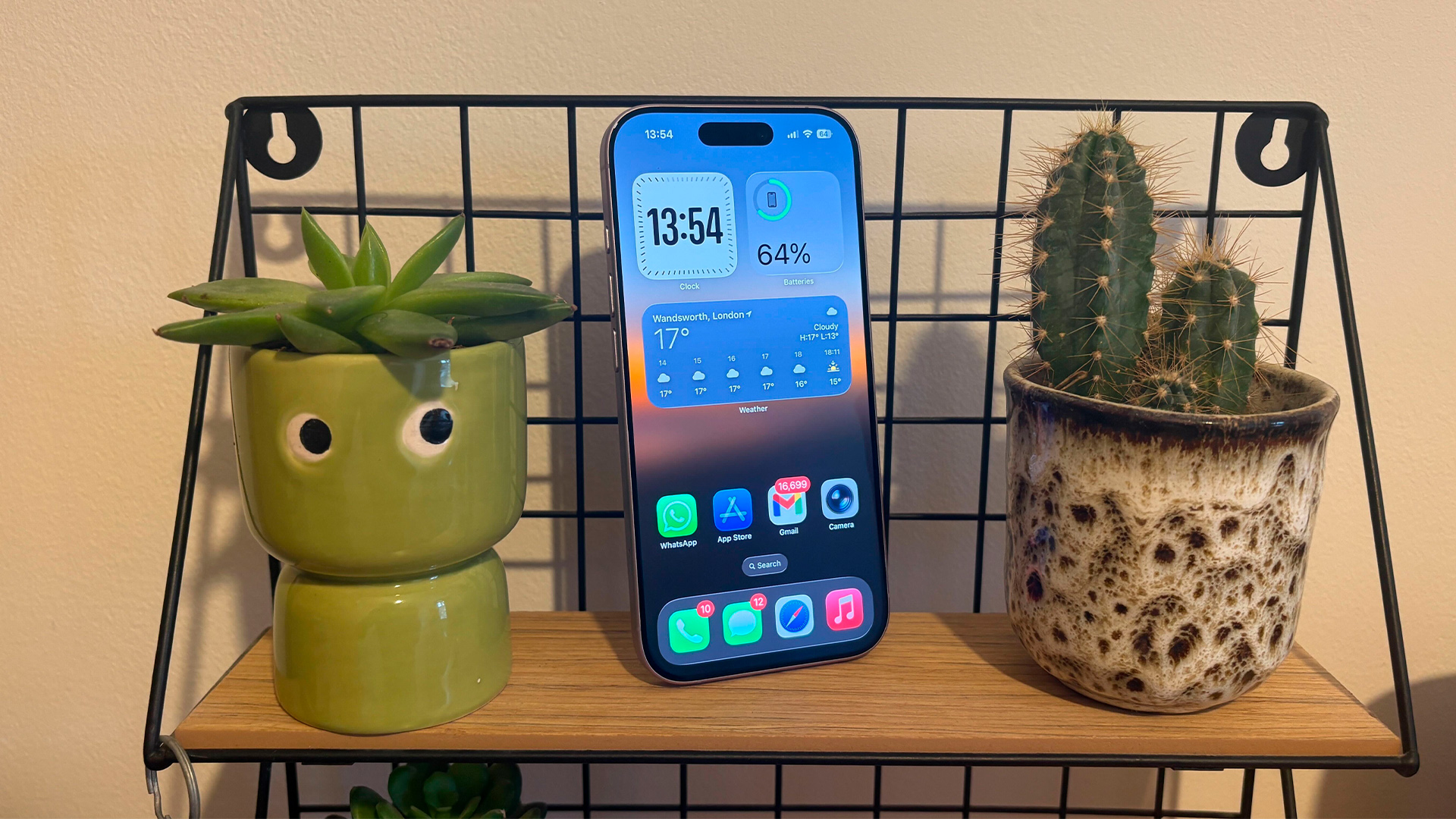 Apple iPhone 17 smartphone upright on shelf between two plantpots, on screen is weather app and time