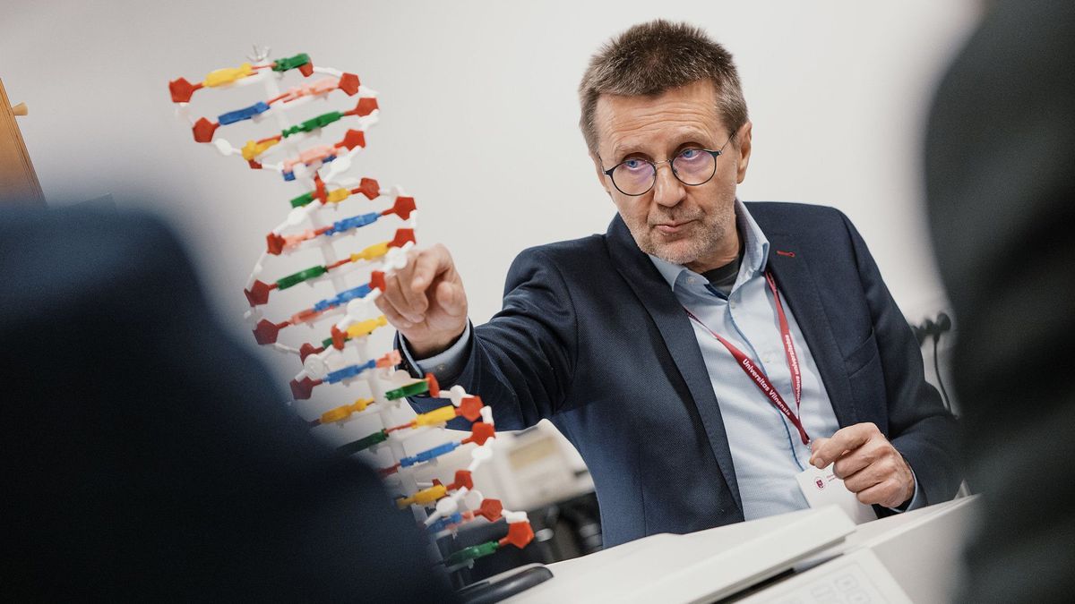 Virginijus Šikšnys (an older man with short brown hair and round glasses wearing a suit jacket) gestures toward a 3D model of DNA on a table