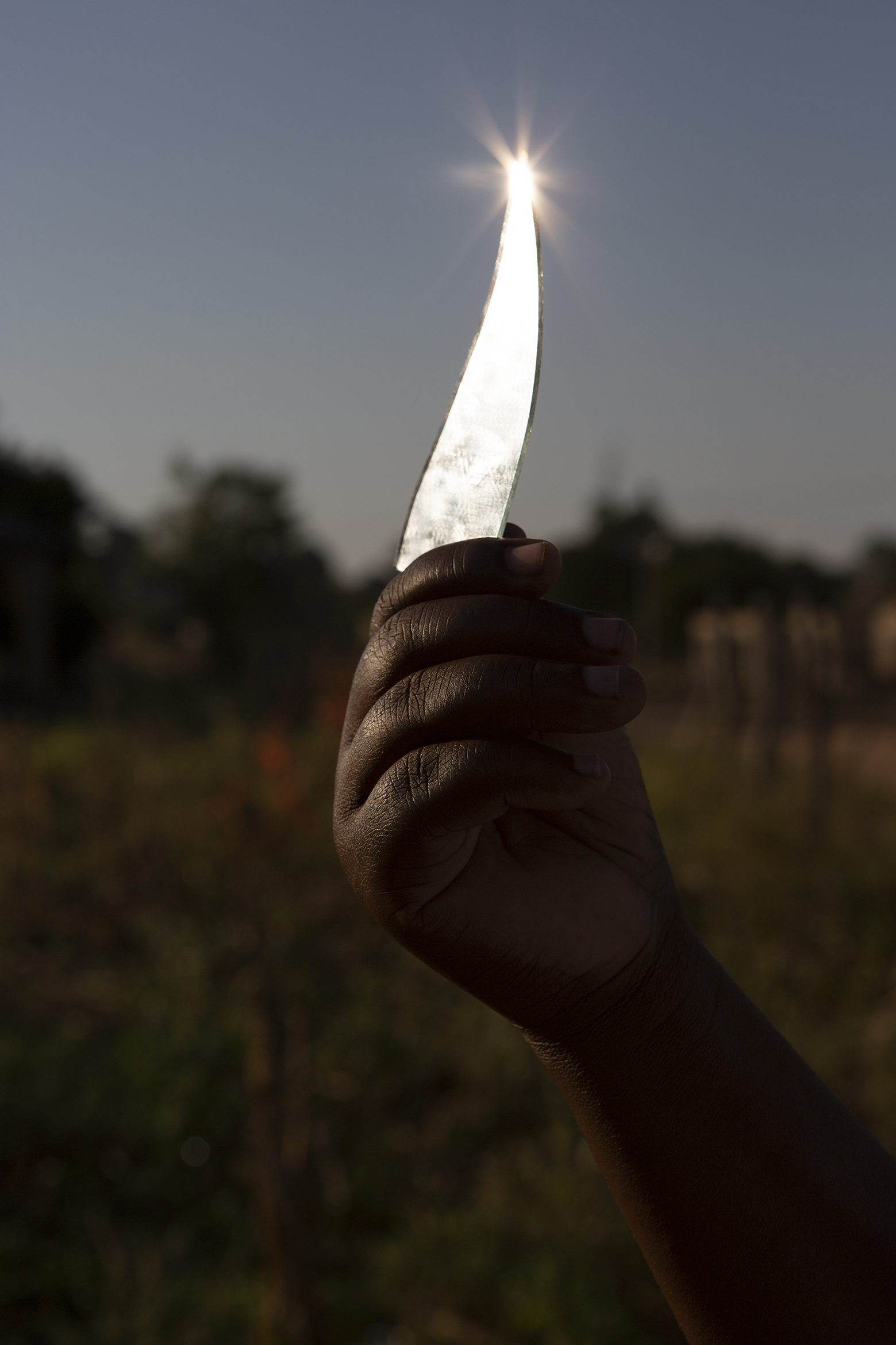 A hand holds a curved glass shard against the sky, capturing sunlight at its tip, creating a bright starburst effect. The background is a blurred landscape