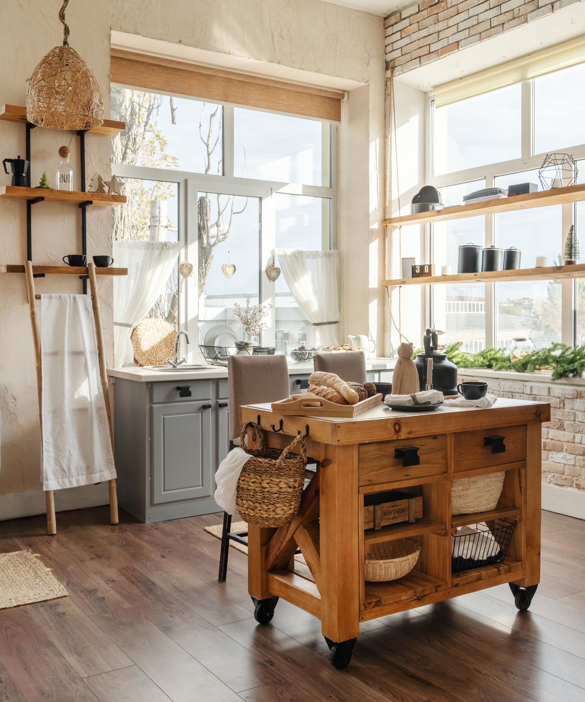 Vertical side view of stylish sunny kitchen interior with kitchenware, home decor and accessories, wooden table with homemade pastry food against cupboards and windows