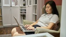 woman slouching in an armchair working on a laptop in her lap. she's in a home setting with a view a white corner unit behind her
