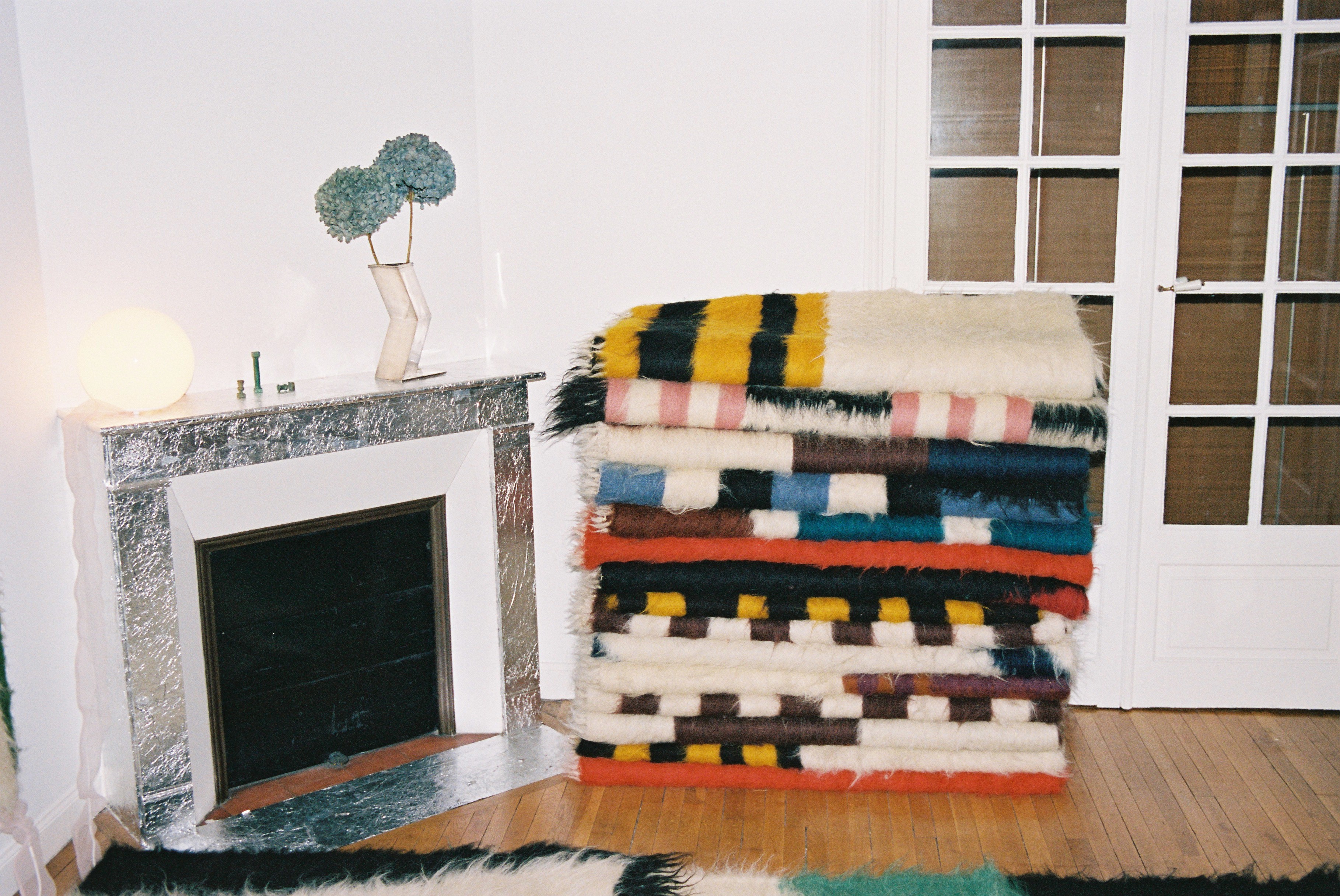 A series of geometric, colorful rugs are stacked on top of each other in a brightly lit living room with a chrome-framed fireplace, a round table lamp, and flowers.