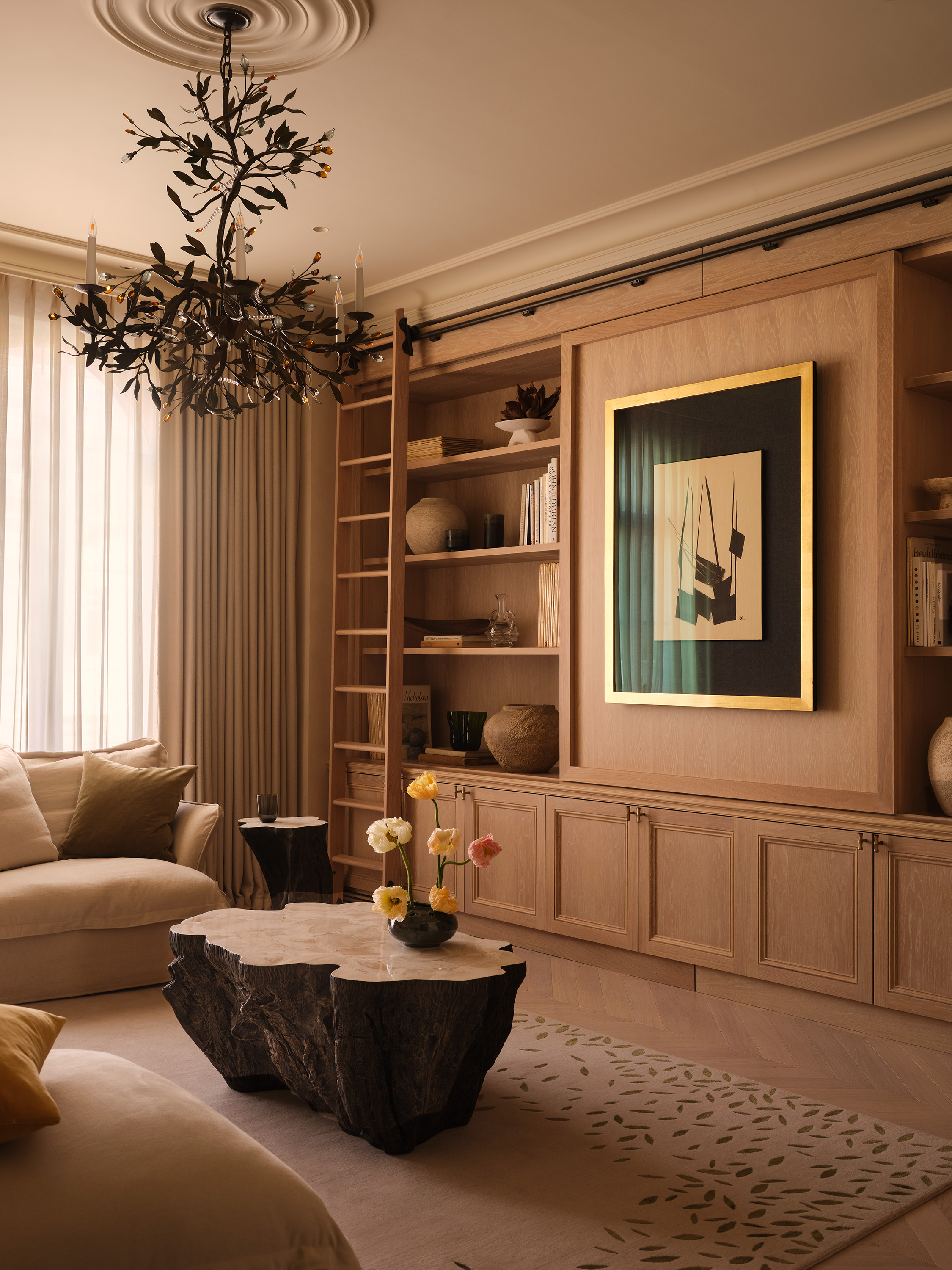 A modern living room with built in storage along the back wall with open decorative shelves either side of artwork in the middle with an abstract coffee table that has colorful flowers in a short round vase underneath a leaf-like chandelier
