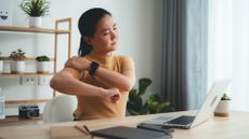 A woman sits at a desk in front of an open laptop with a slightly pained expression. She is holding her right shoulder with her left hand and stretching it out to the side by lifting her elbow.