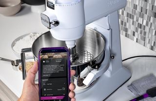 En hand håller upp en mobil som kör den tillhörande appen för Ge Profiles Smart Mixer, med köksapparaten på en bänk i bakgrunden.
