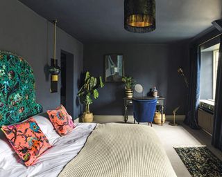 Eclectic and dramatic bedroom with dark blue walls and ceiling, wallpapered feature wall and double bed with white bedding and bright cushions. feature headboard with dramatic and colourful fabric