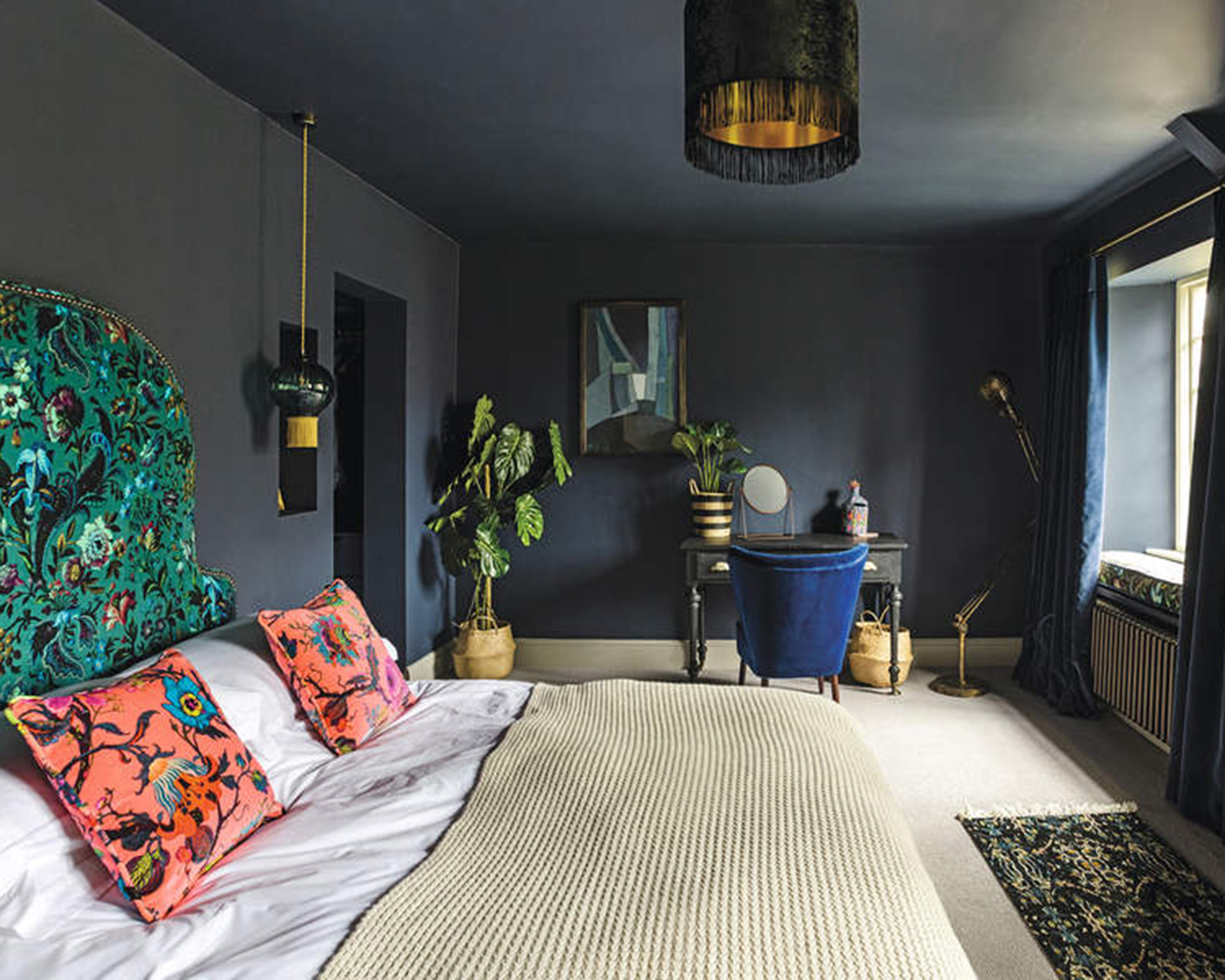 Eclectic and dramatic bedroom with dark blue walls and ceiling, wallpapered feature wall and double bed with white bedding and bright cushions. feature headboard with dramatic and colourful fabric