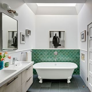 White bathroom with green scalloped tiles on half height wall and small white bath on grey tiled floor