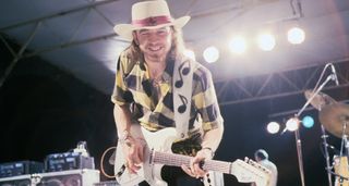 Stevie Ray Vaughan smiles as he plays on a brightly lit stage. He plays a white Strat, with a thick white strap with musical notes and wears a wide-brimmed white hat.