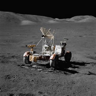Lunar Roving Vehicle pictured on the surface of the moon with nobody in it. It has four wheels and a parasol-like antenna sticking out of the top.