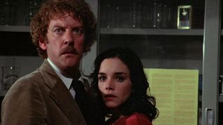 A man with a brown suede jacket, matching his curly hair and moustache, looks startled as he stands right next to a woman with long black hair, wearing a red top