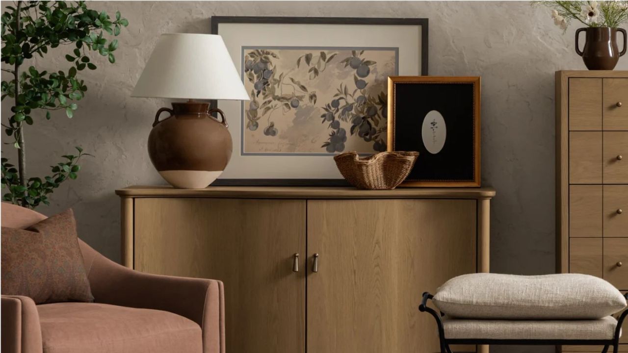 A living room vignette: a lamp on a console table, beside a pink armchair and a cream stool, against a gray wall.