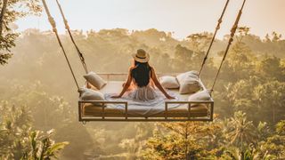 A woman taking part in sleep tourism by sitting on a bed that&#039;s suspended above a rainforest