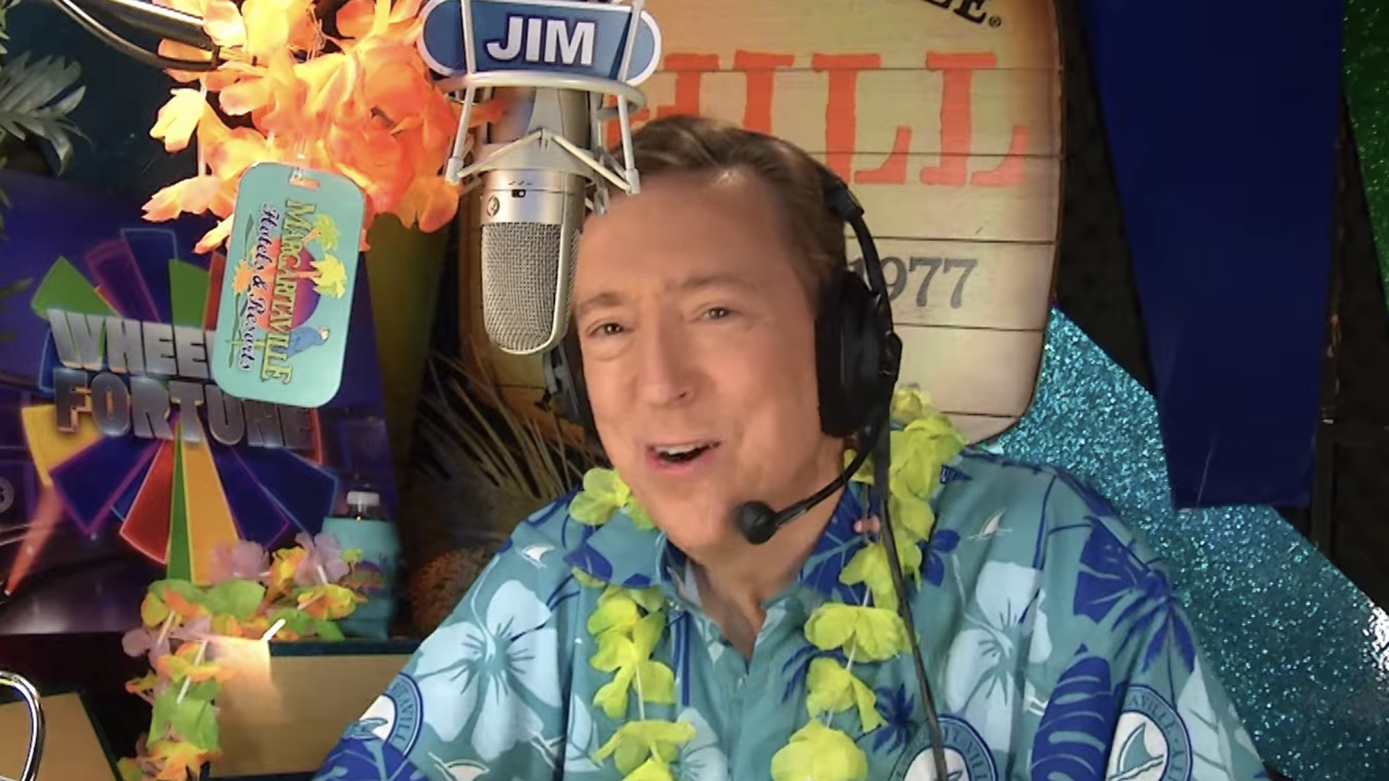 Wheel of Fortune announcer Jim Thornton sitting in booth behind microphone wearing Hawaiian shirt and leis