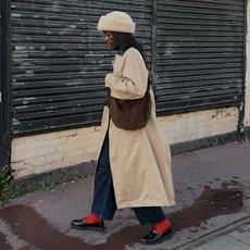 influencer wears trench coat, jeans, red socks with loafers, hat, and cross body brown espresso suede bag