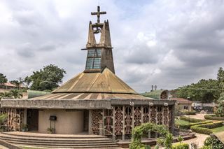 Daytime image of Nwoko’s first commission, in 1970, a chapel for the Dominican Institute in the Nigerian city of Ibadan, green hedges and shrubs, trees, stone steps up to the chapel, surrounding landscape and buildings, cloudy sky