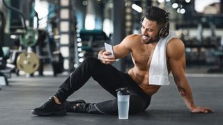 Man sitting on the floor of a. gym post-workout holding his phone smiling