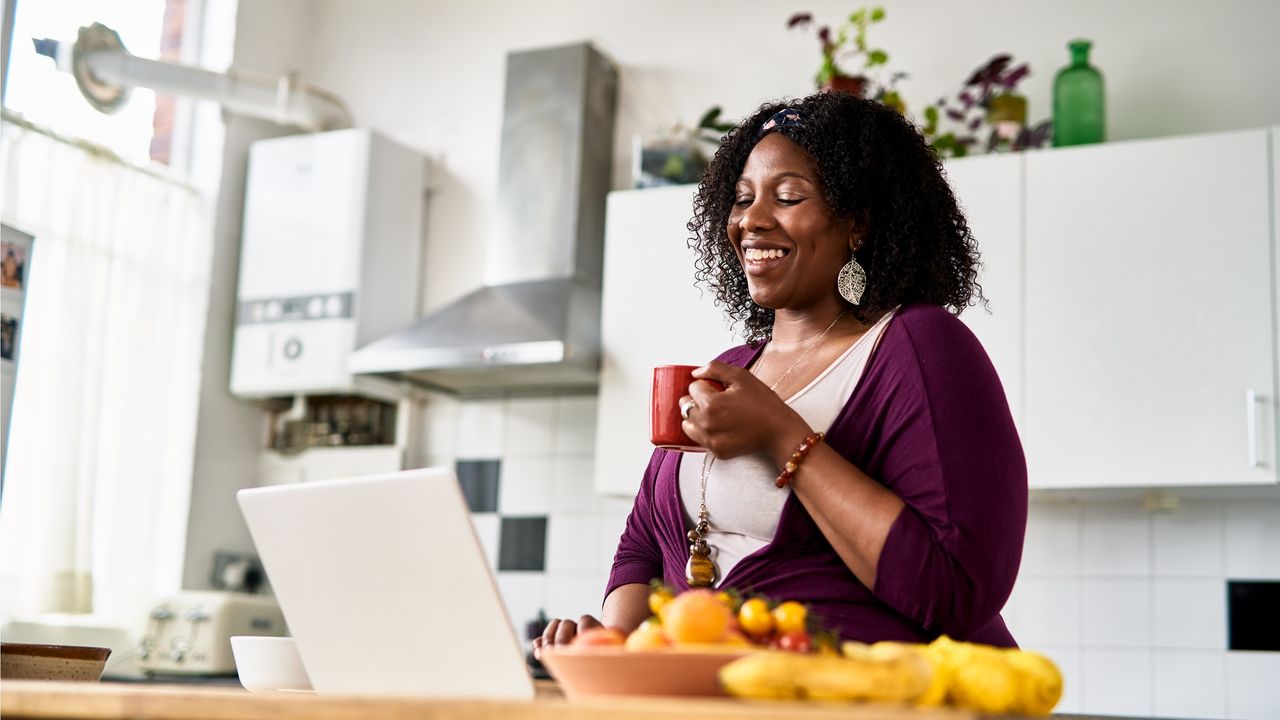 A very happy woman sits in her kitchen looking at her laptop.