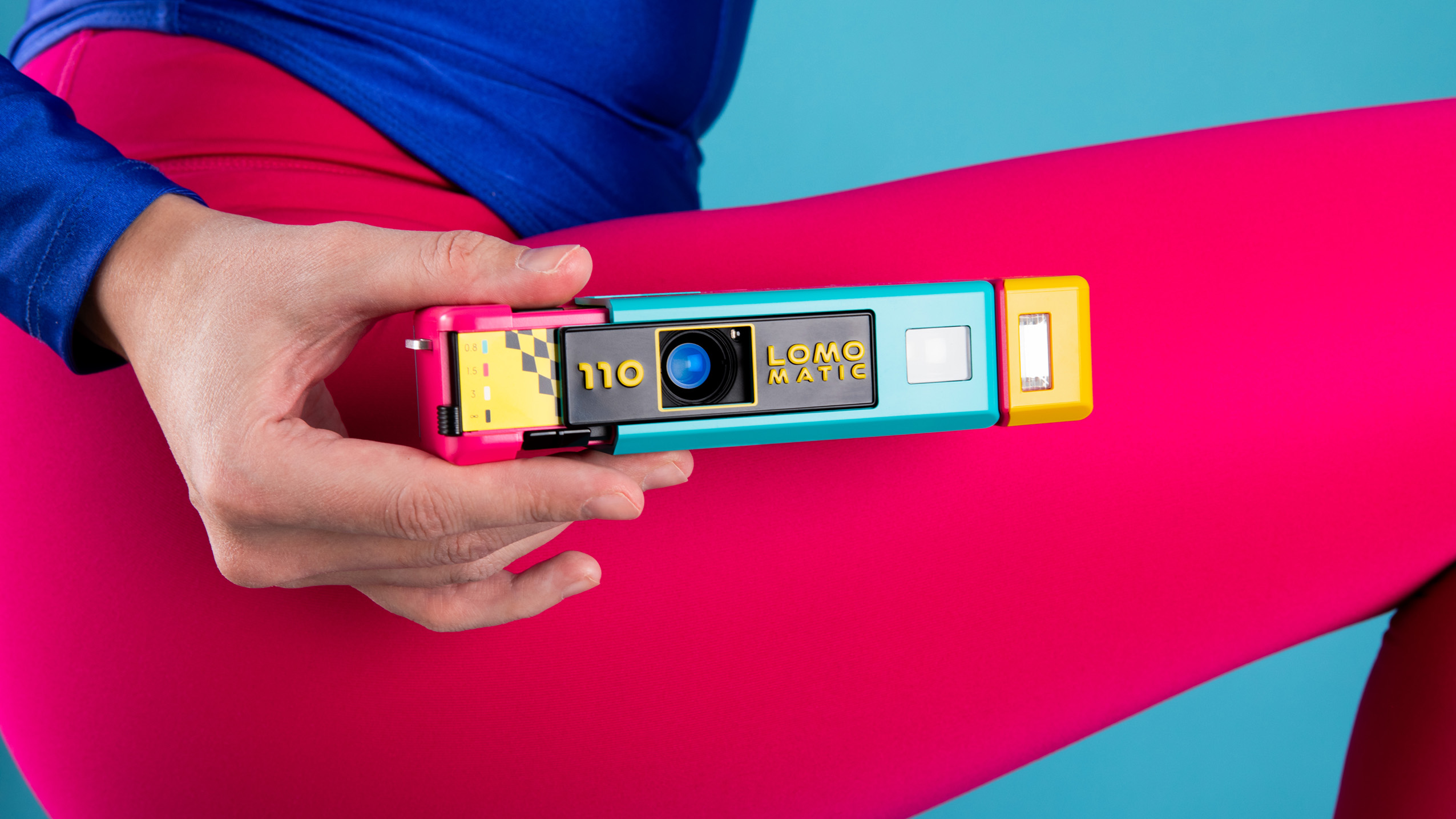Hand holding a colorful Lomomatic 110 film camera against bright pink leggings and a blue background