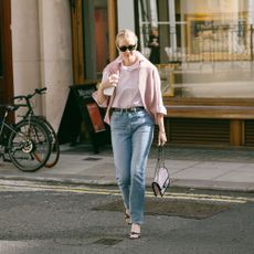 Kristen walks on London street in pink button down, jeans, heels, handbag, and a sweater over her shoulders. 