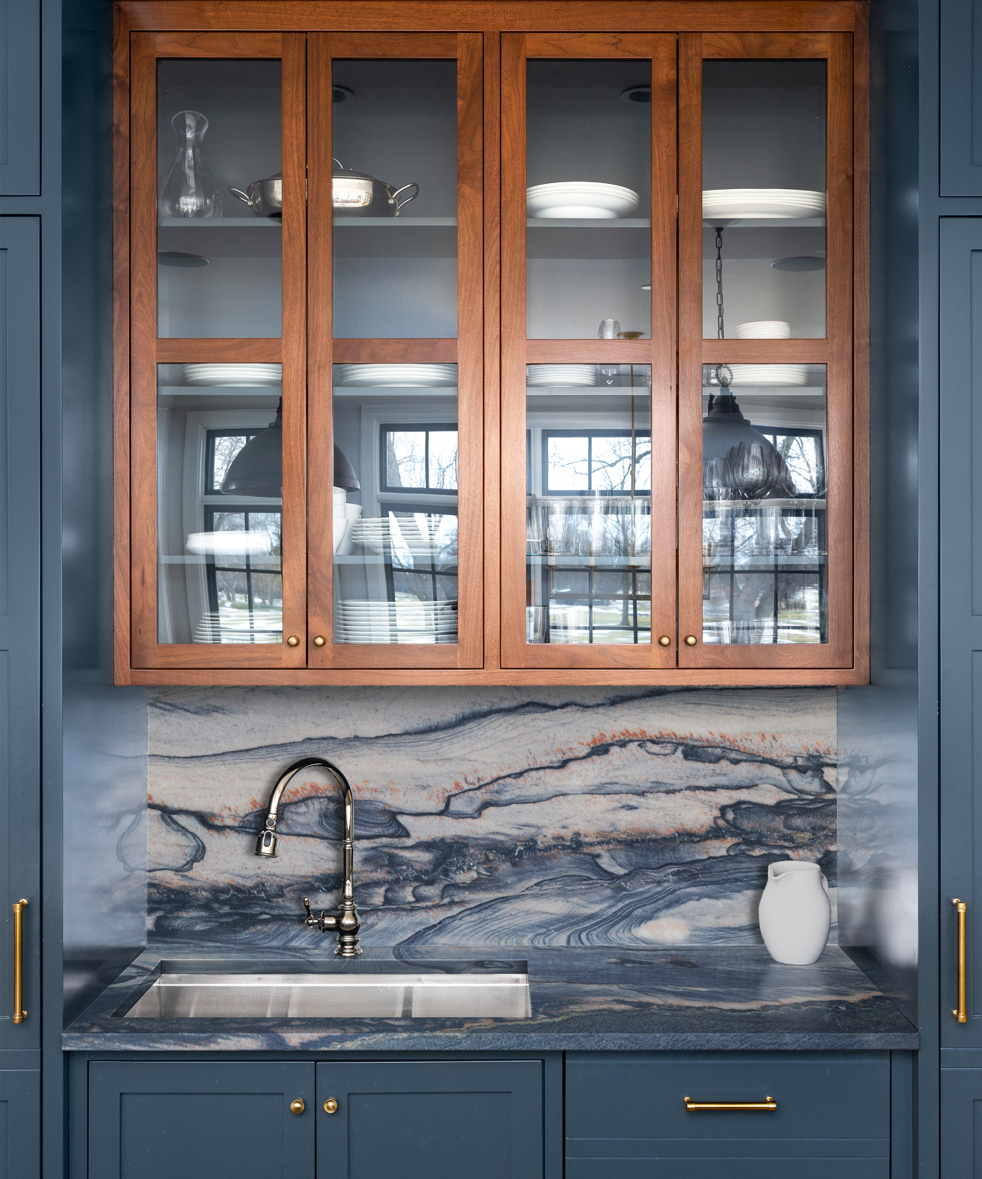 Kitchen sink area with dark blue veined marbled worktop and splashback with glass fronted wooden cabinet above and navy cabinets below