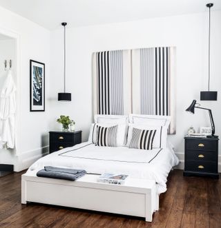 White bedroom with striped headboard, hardwood floor, black matching side tables, black matching low hung pendants, wooden ottoman, artwork