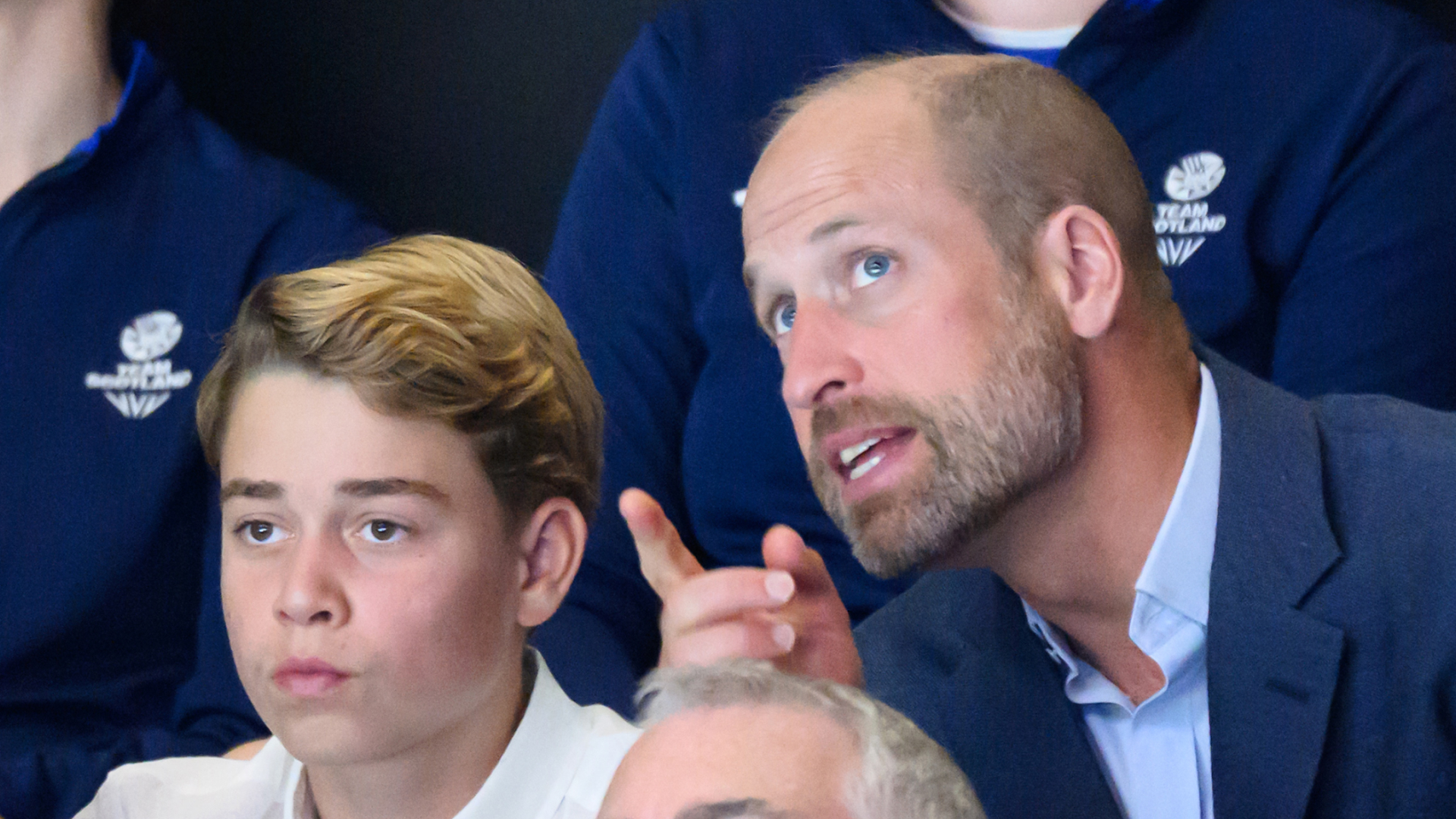 Prince George looks serious and wears a white shirt while his dad Prince William wears a navy suit jacket and points at something, he has a beard but no hair