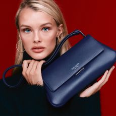 Model holding a deep blue Kate Spade handbag on a burgundy background 