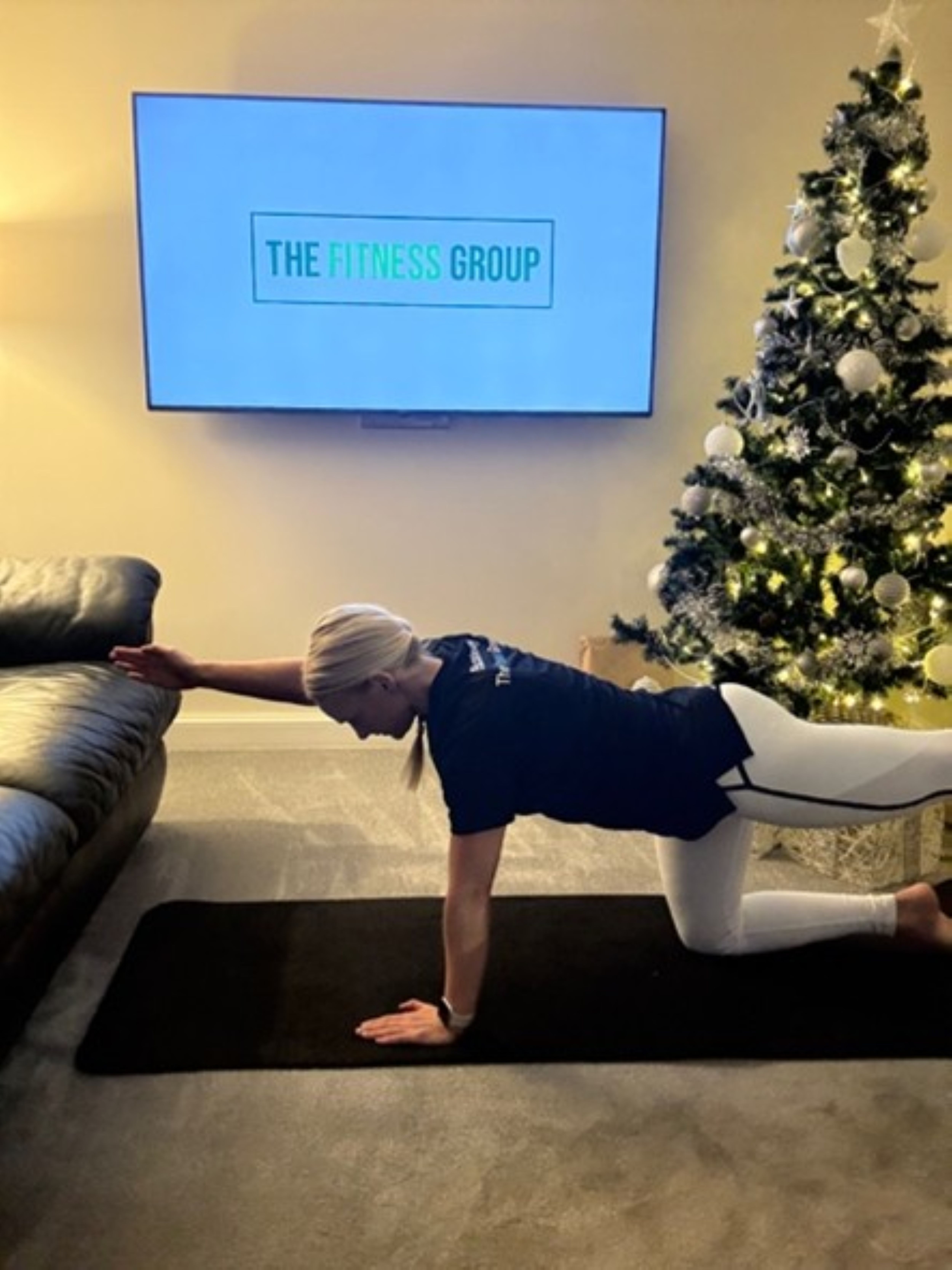 Woman demonstrates a Pilates exercise between a sofa and a Christmas tree. A mounted TV in the background reads "The Fitness Group"