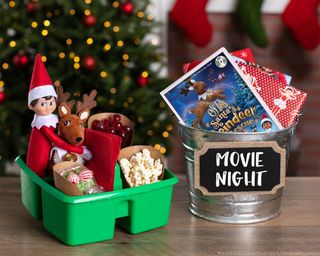 An elf in a green caddy container with plush reindeer, grapes, popcorn and confectionery with movie night bucket containing DVDs