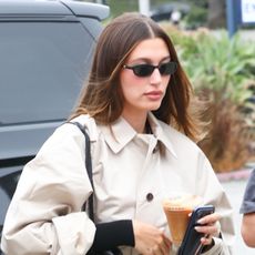 Hailey Bieber wearing a trench coat, black flare pants, and plain black leather handbag at the DMV
