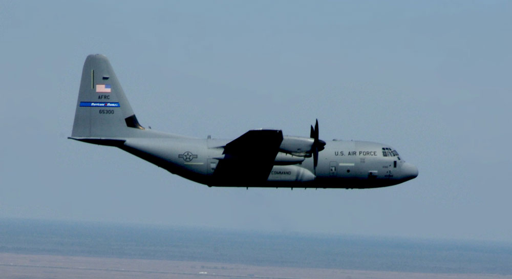 A WC-130J "Hurricane Hunter" aircraft flies over the coastline.