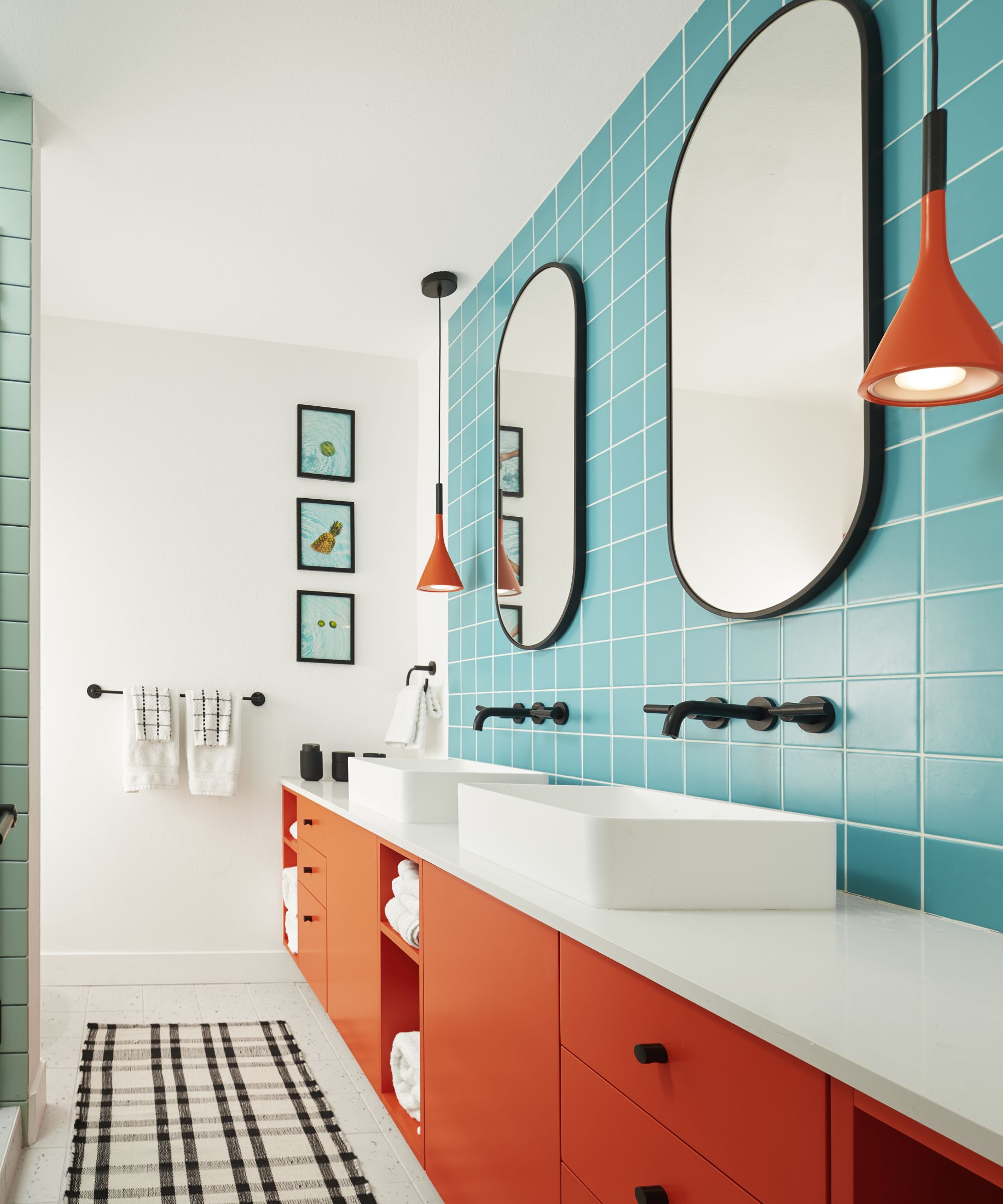 Bathroom with white walls, blue tiled backsplash with round black mirrors and his and hers sink, with orange painted shelving underneath and low orange pendant lights on either side