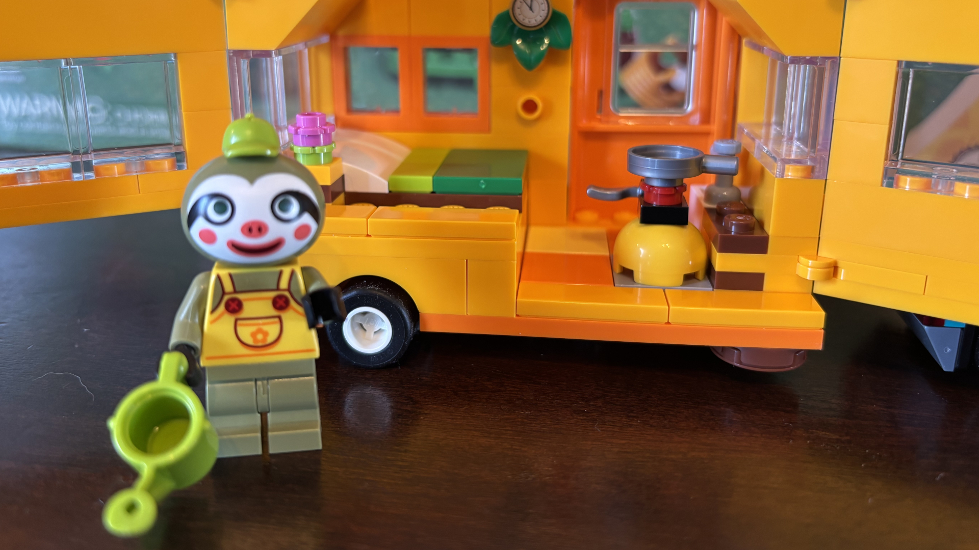 Lego Leif&#039;s Caravan with a Leif minifigure in front of it holding a watering can, all on a wooden surface